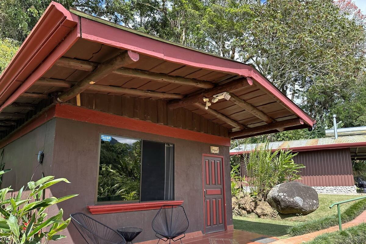 Un pequeño edificio de techo rojo con una ventana, rodeado de follaje.