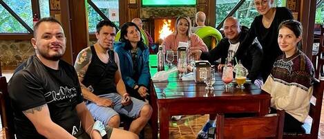 Um grupo de pessoas sentadas em torno de uma mesa de madeira em um quarto com parede de pedra e uma tela de televisão.