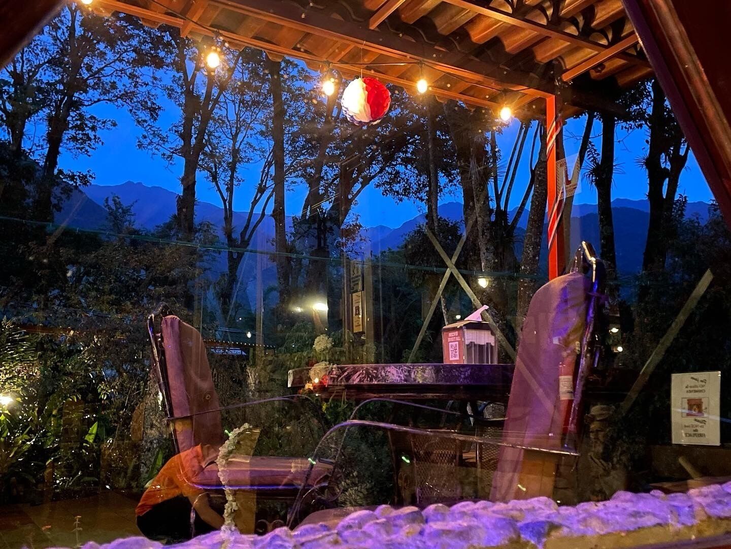 Un rincón al aire libre acogedor con dos sillas y una mesa, rodeado de abundante vegetación y vistas a montañas.