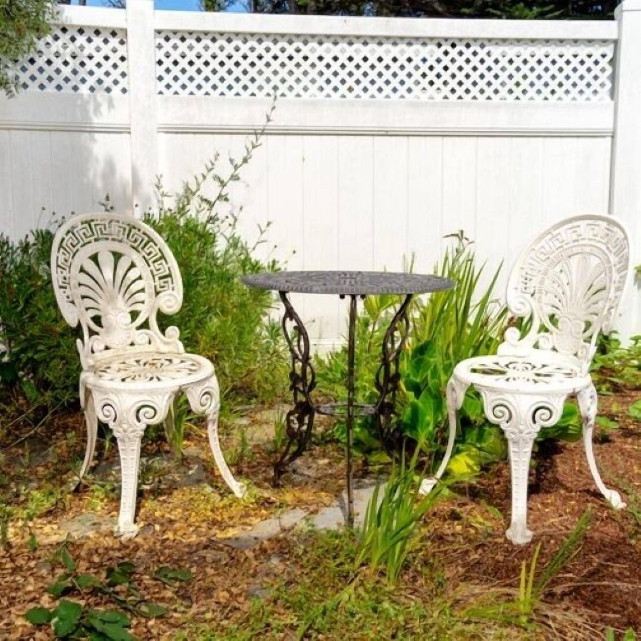 Deux chaises en métal blanc, richement décorées, et une petite table ronde, le tout installé dans un jardin.