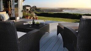 A patio with wicker furniture, a table with a bottle and glasses, and a view of the ocean and hills. - Agate Cove Inn (Mendocino)
