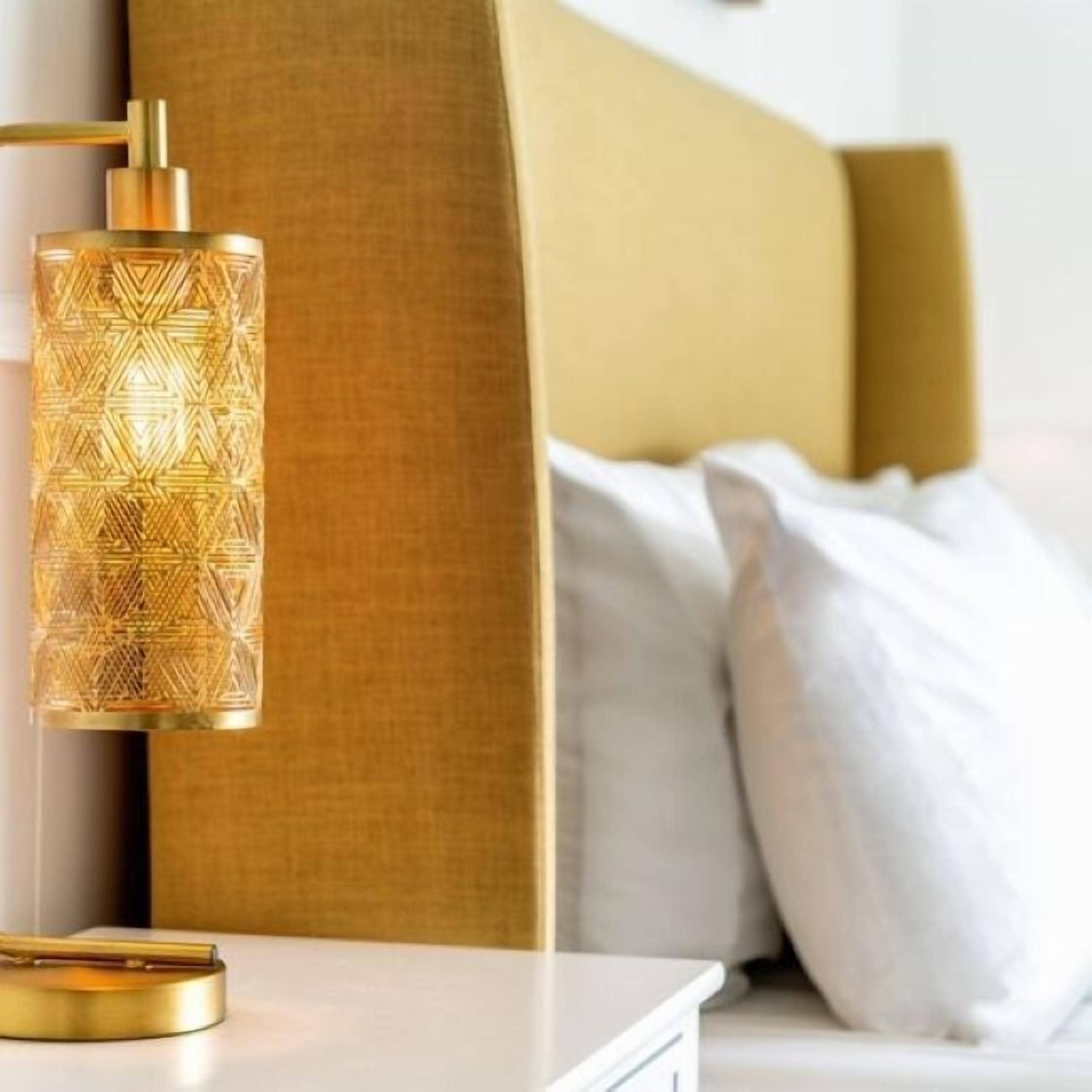 Una cama moderna con cabecera dorada, una mesita de noche con lámpara dorada y ropa de cama blanca.
