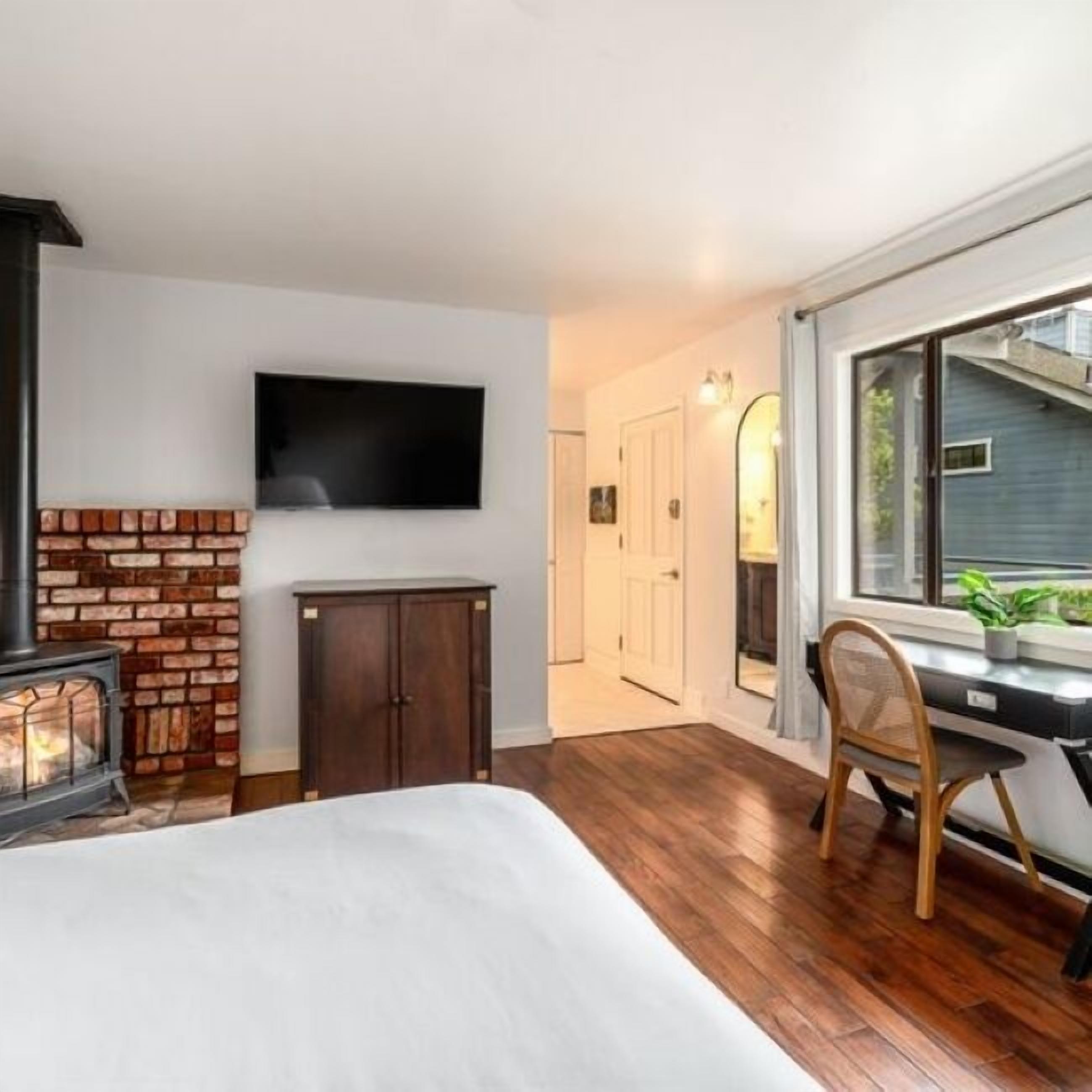 A bedroom with a fireplace, a TV mounted on the wall, a desk with a chair, and a window with a view of the outside.