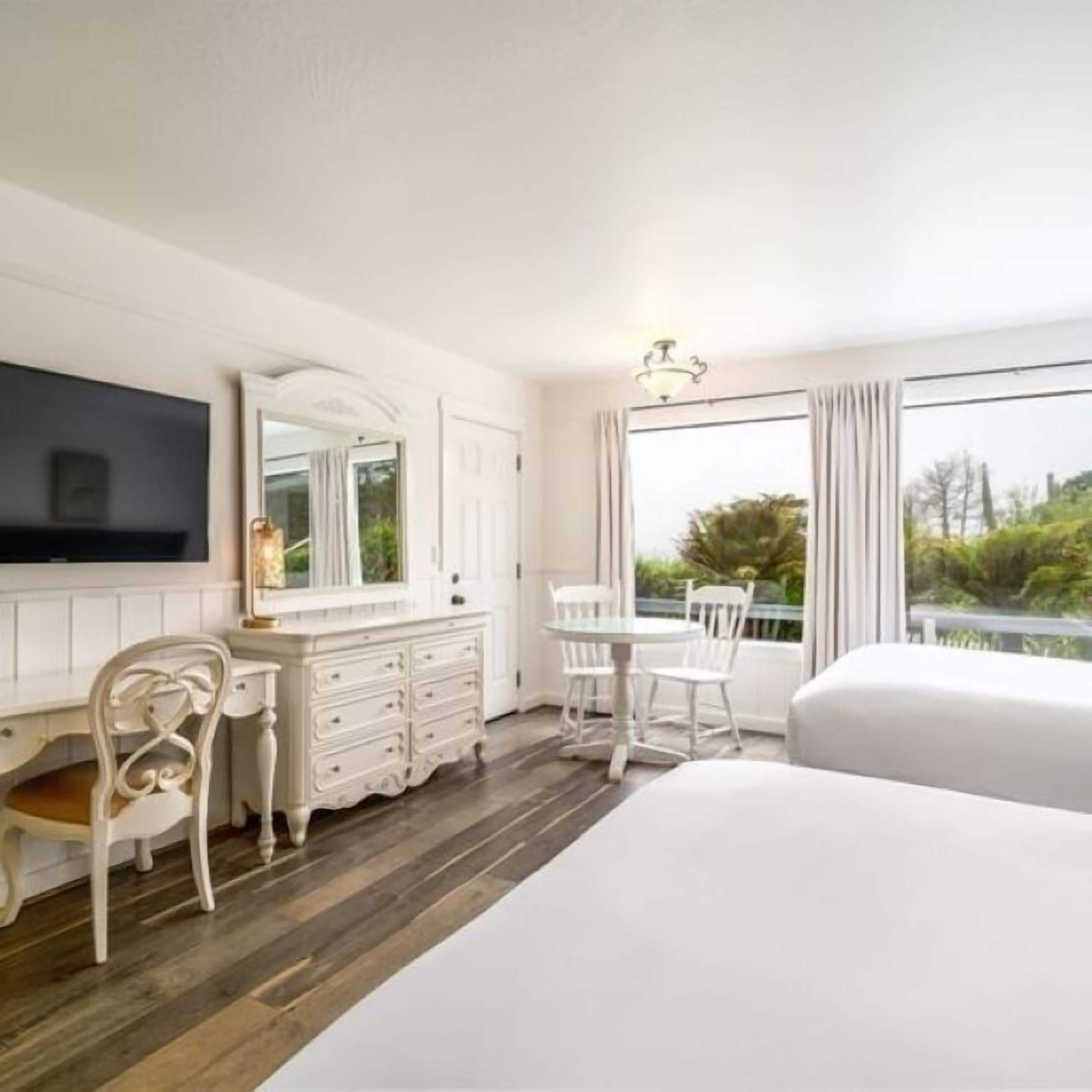 A hotel room with a large bed, a desk with a chair, a dresser, and a TV mounted on the wall. There is a window with a view of greenery outside.