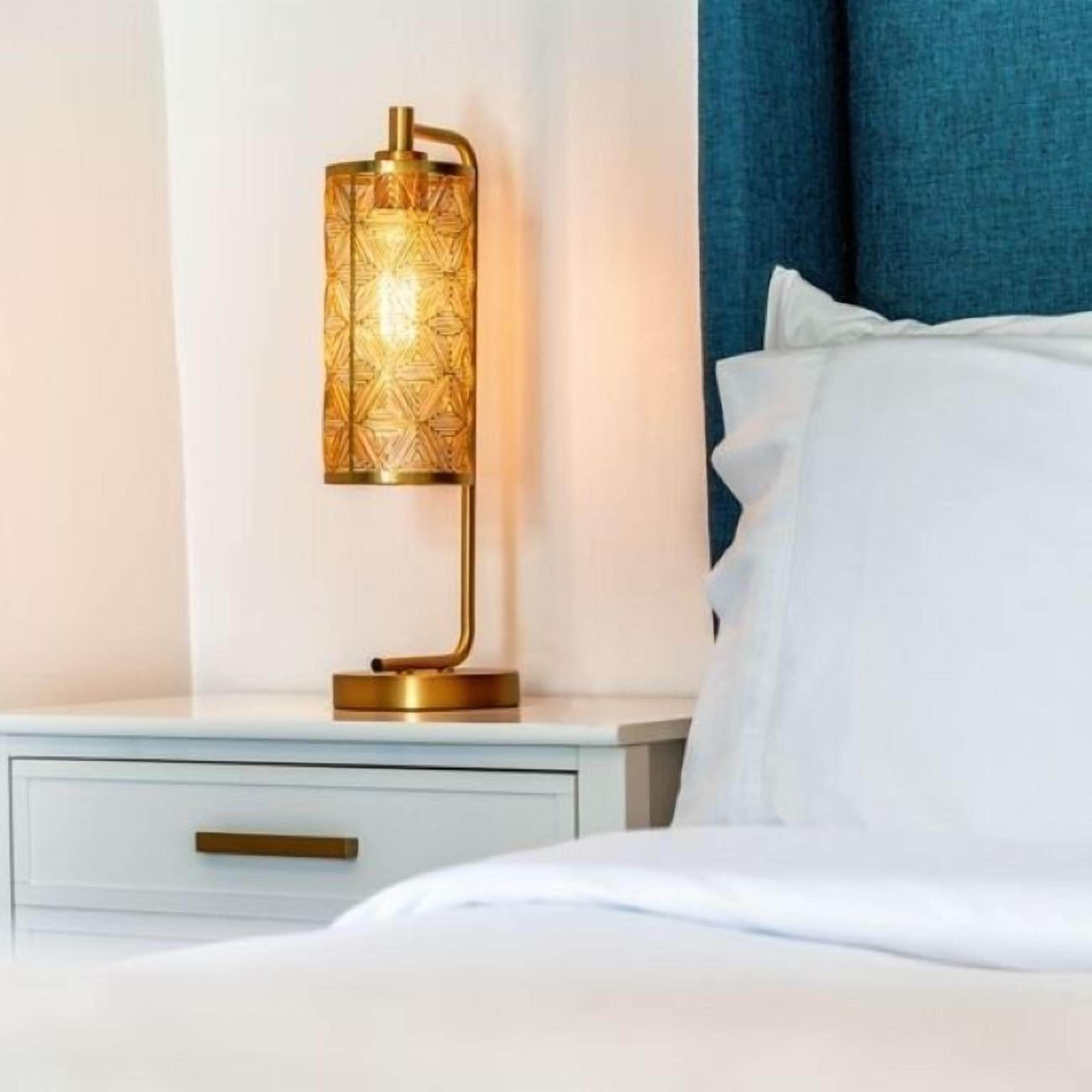 A bed with white bedding, a bedside table with a gold lamp, and a blue curtain in the background.