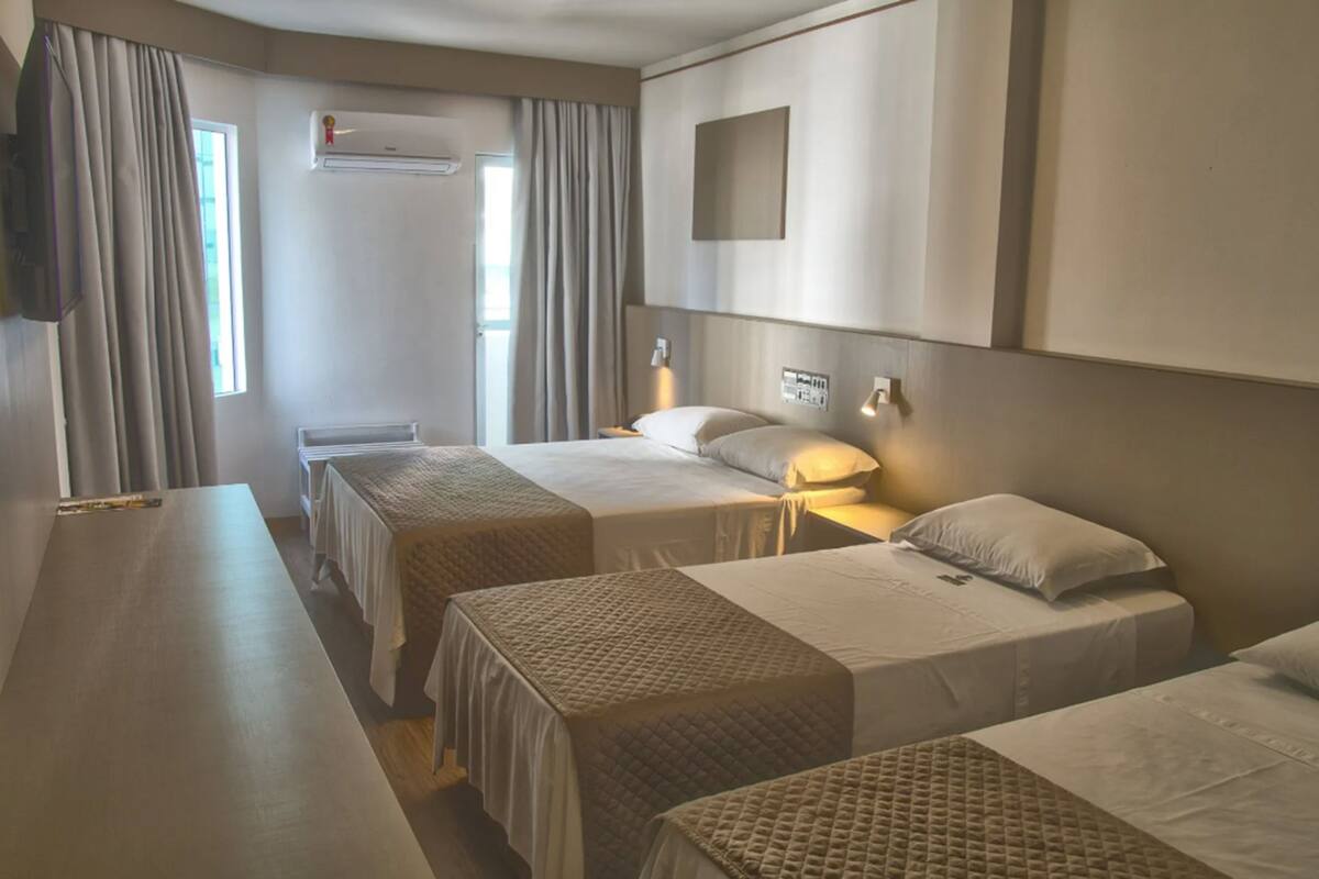 Ein Hotelzimmer mit zwei Einzelbetten, einem Schreibtisch, einem Stuhl, einer Klimaanlage und einem an der Wand montierten Fernseher.