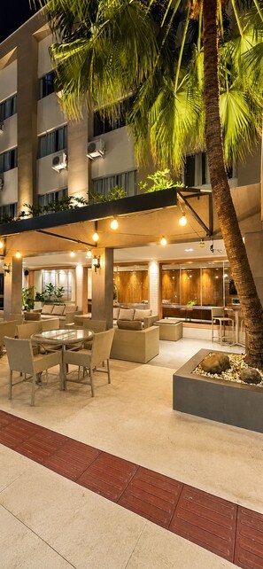 An outdoor lounge area with seating and a dining table, surrounded by palm trees and a building with multiple floors. - Hotel Gracher Praia (Balneário Camboriú)
