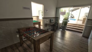 A room with a foosball table, a small pool table, a desk with a computer, and a potted plant.