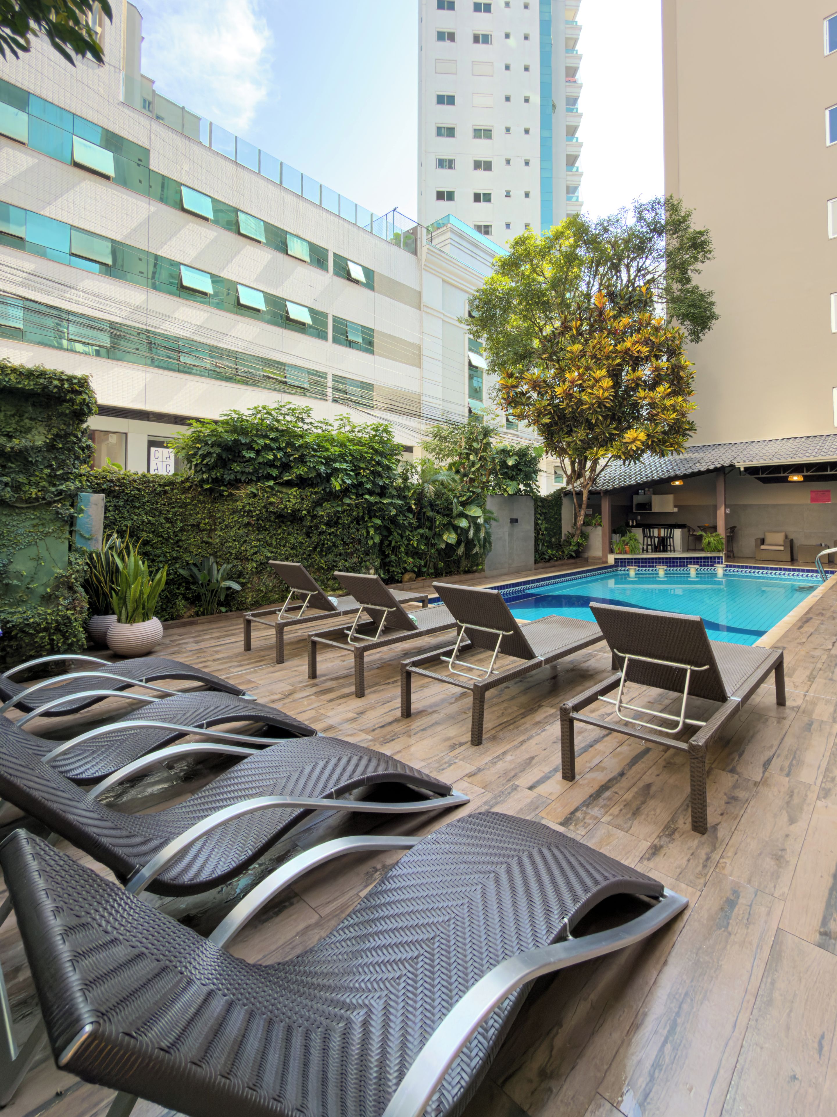 Área de piscina no terraço com espreguiçadeiras e espaço para refeições, cercada por áreas verdes e edifícios modernos.