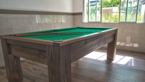A pool table in a room with wooden flooring and a window with a view of greenery outside.