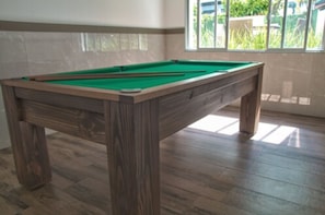 A pool table in a room with wooden flooring and a window with a view of greenery outside. - Hotel Gracher Praia (Balneário Camboriú)