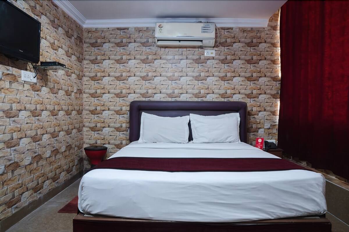 A hotel room with a brick-patterned wall, a bed with white bedding, a red throw blanket, a mounted TV, and an air conditioning unit.
