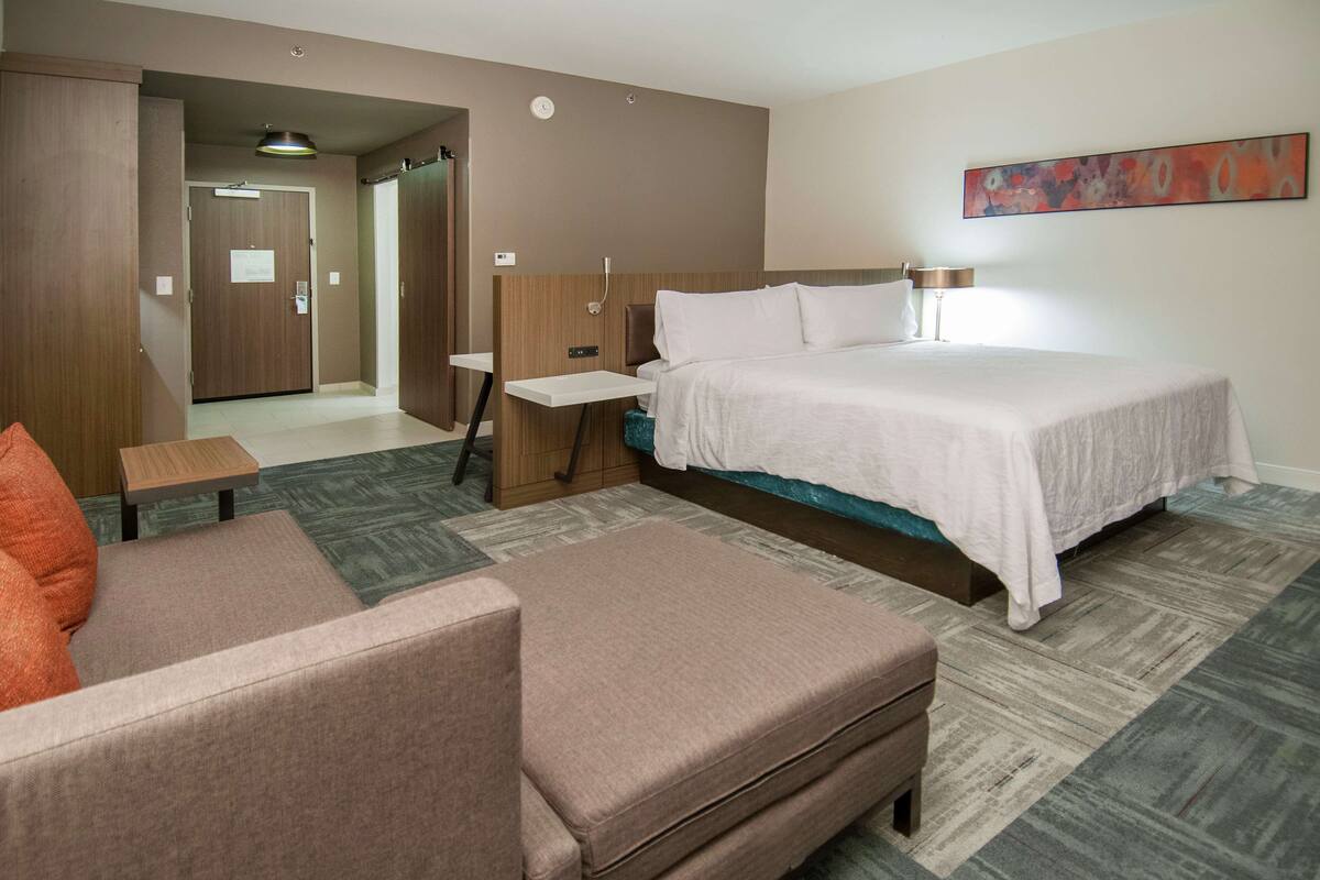 A hotel room with a bed, a desk, a chair, and a sofa. There is a door leading to another room.
