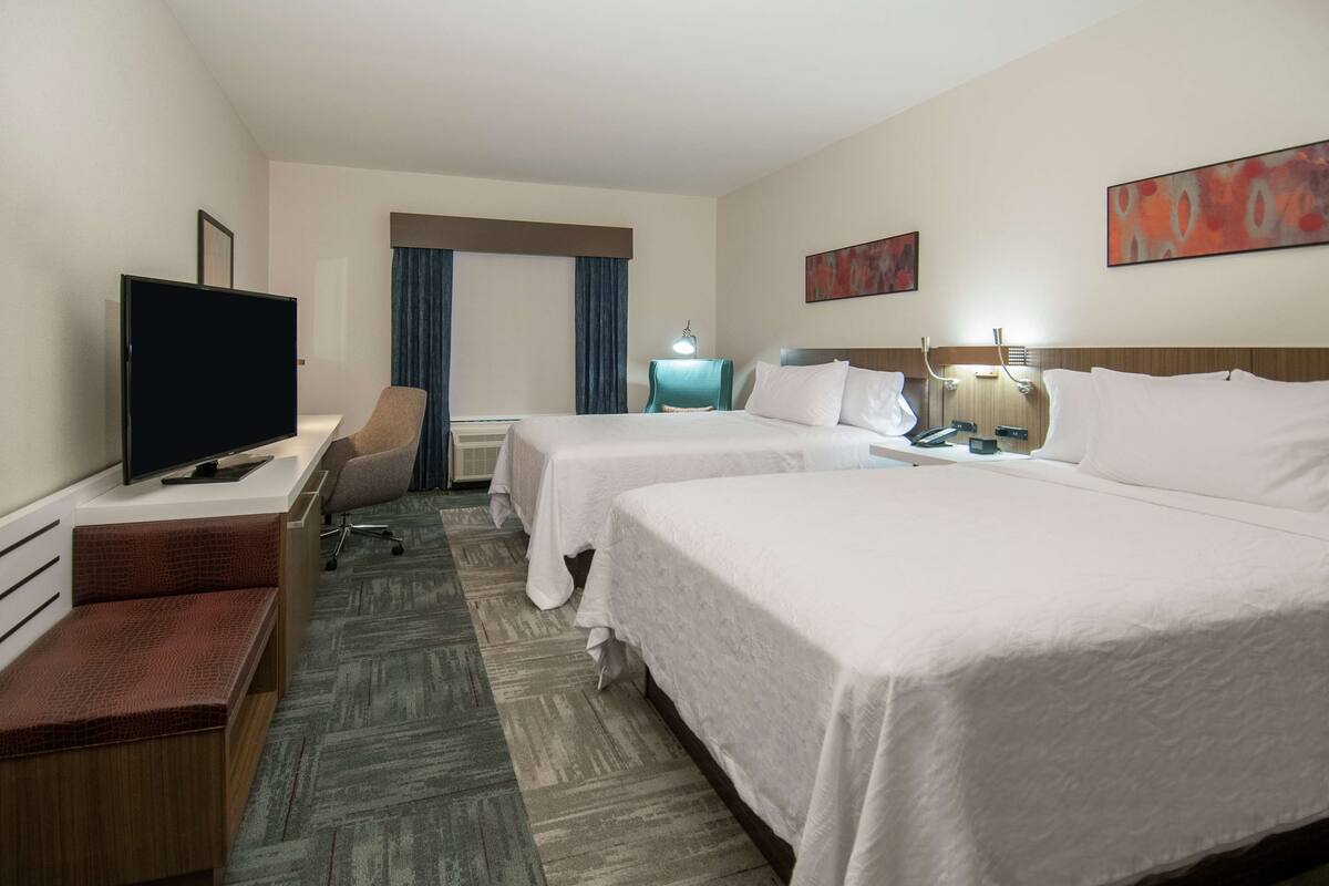 A hotel room with two beds, a desk with a television, a chair, and a bench.