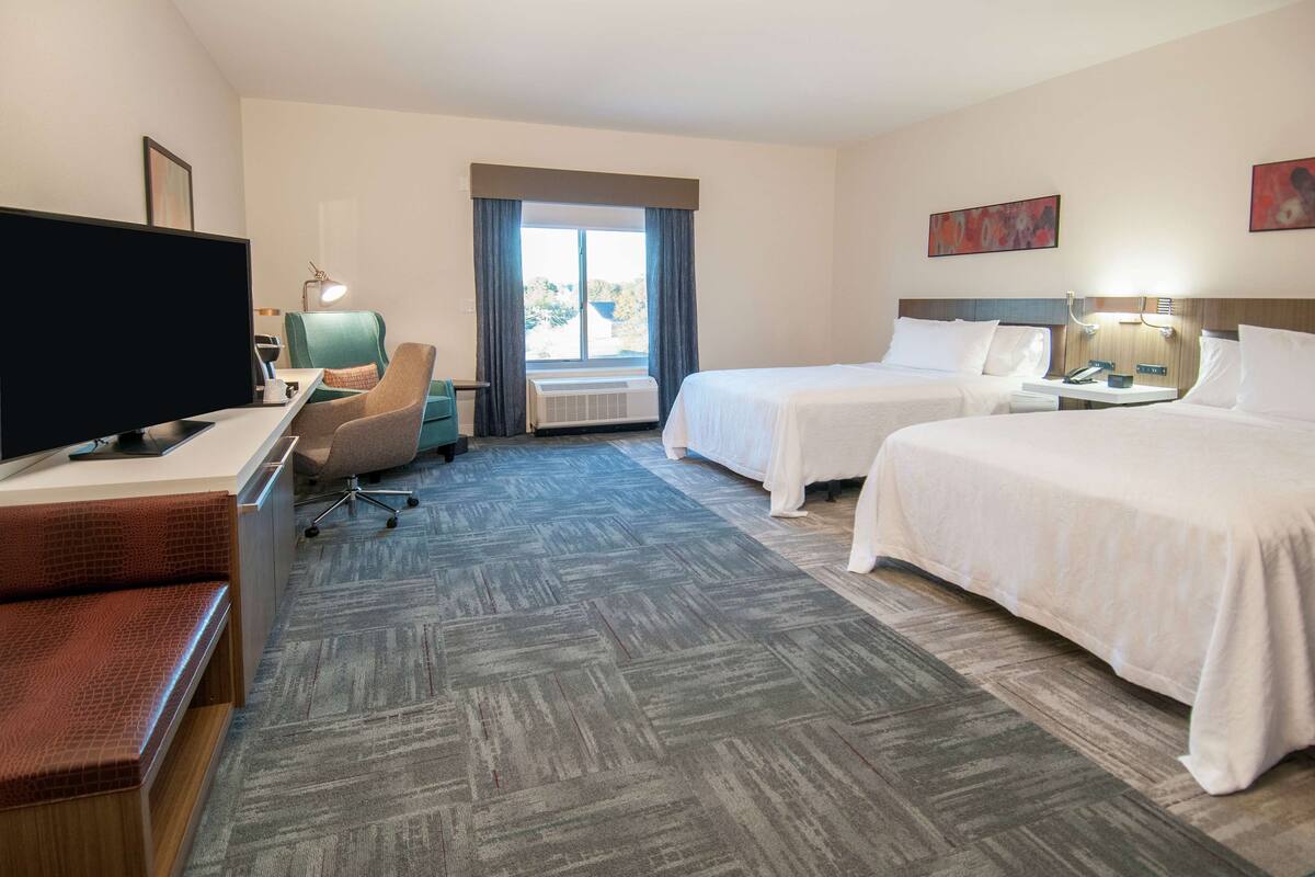 A hotel room with a desk, two beds, a television, and a window with curtains.
