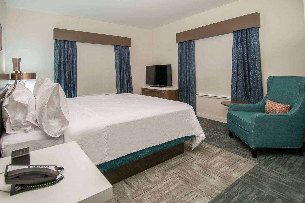 A hotel room with a bed, a desk with a telephone, a television, and a teal armchair.