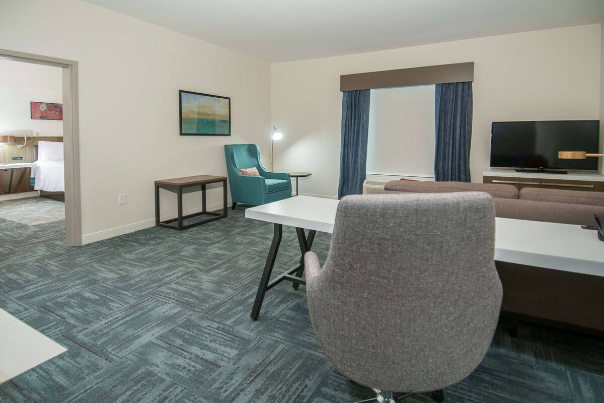 A hotel room with a large window, a flat-screen TV, a sofa, a dining table, and a chair.