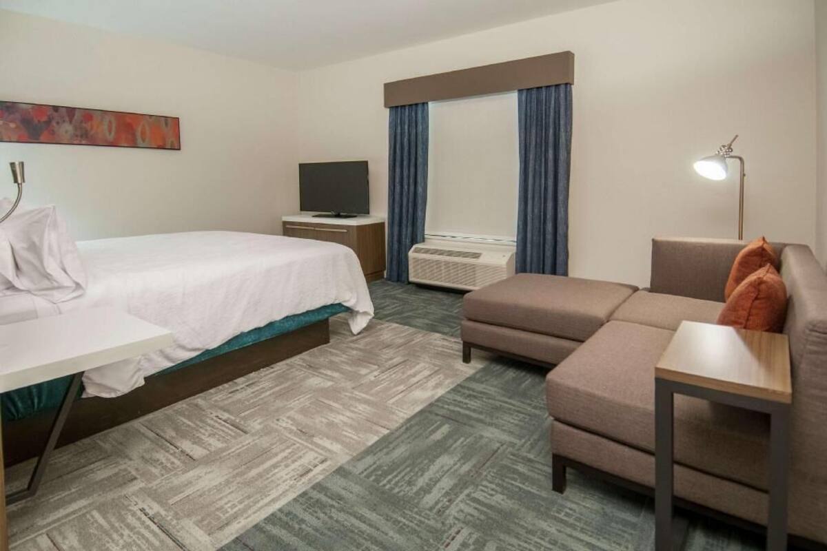 A hotel room with a bed, a sofa, a desk, a TV, and an air conditioning unit.