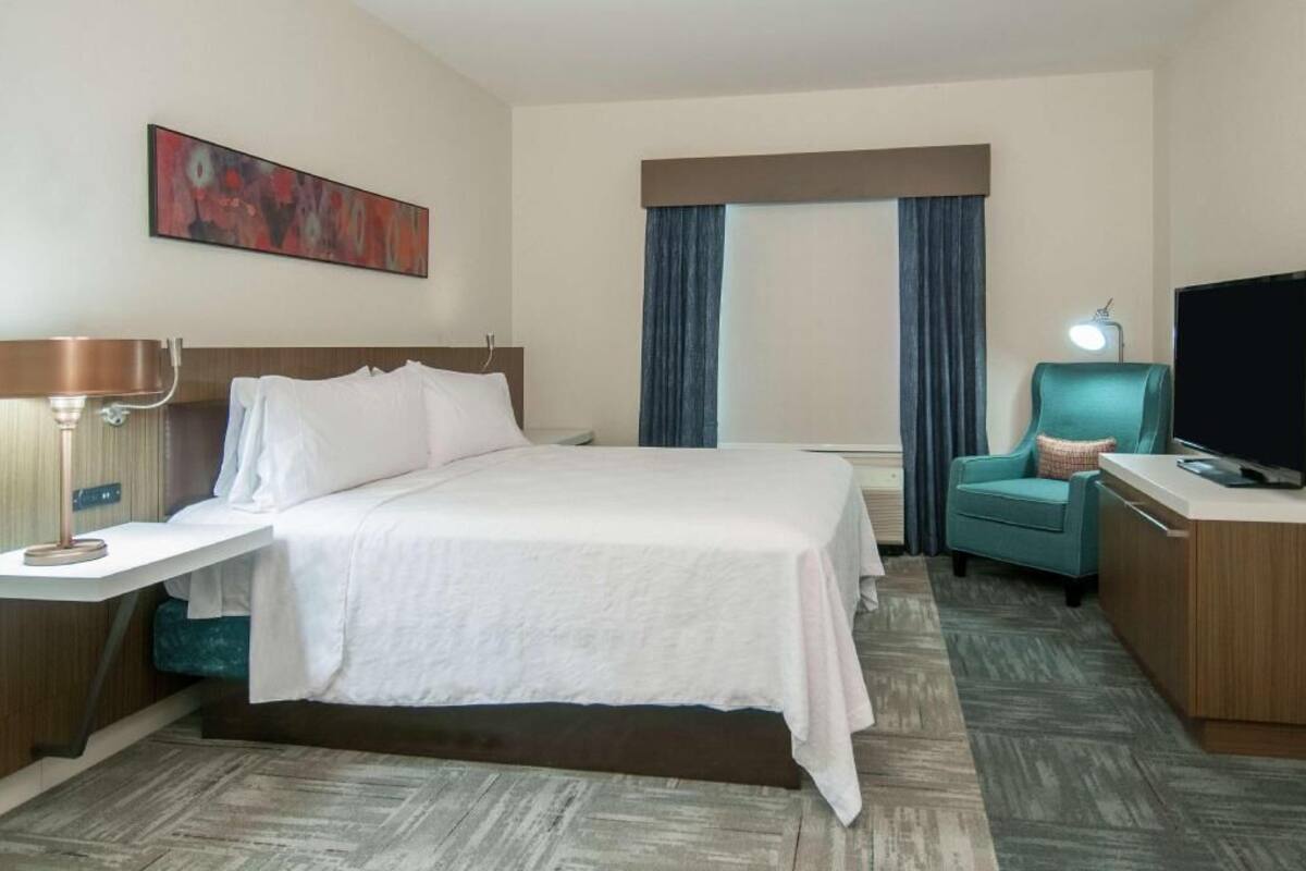 A hotel room with a bed, a desk with a lamp, a TV, a chair, and a window with curtains.