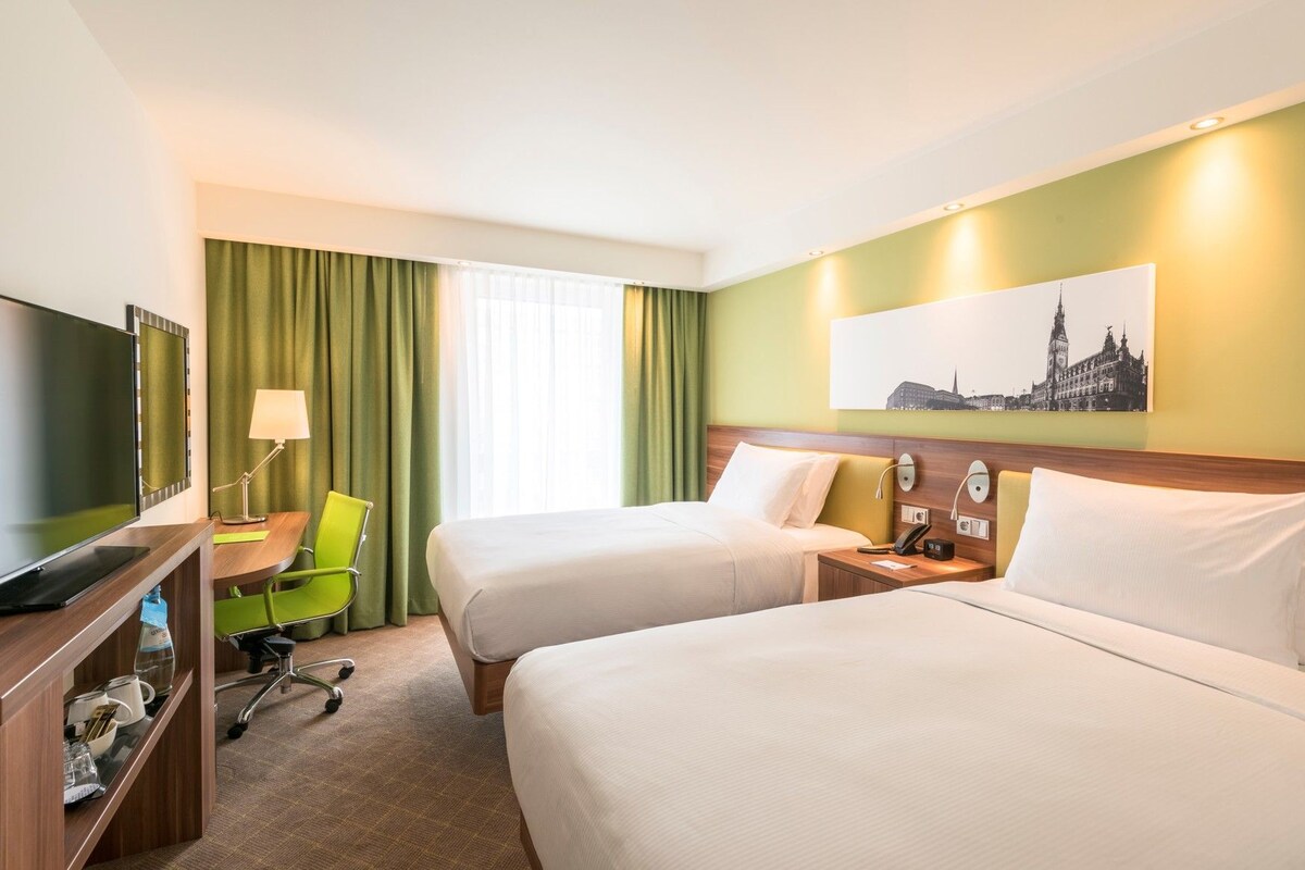 A hotel room with two beds, a desk with a chair, a television, and a painting on the wall.
