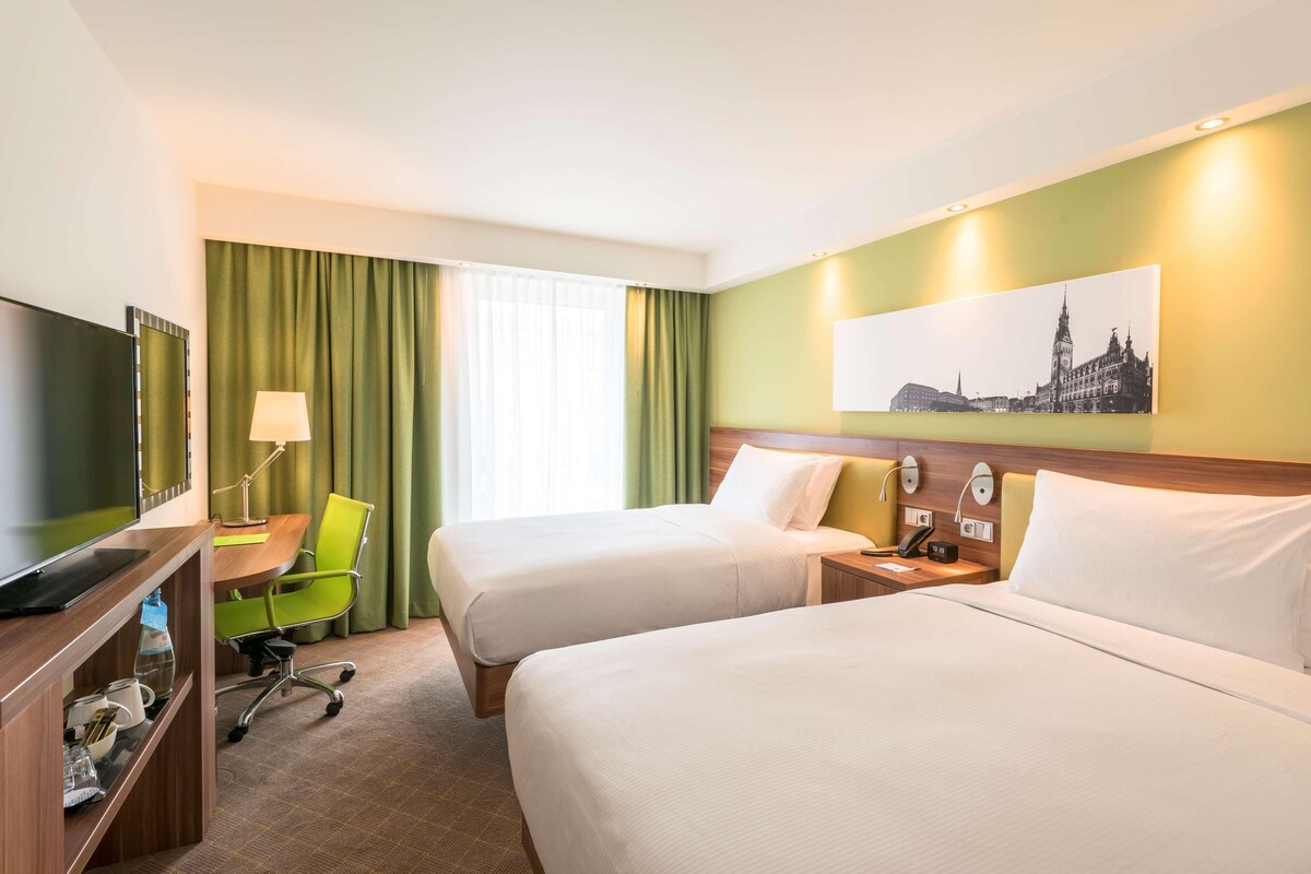 Ein Hotelzimmer mit zwei Betten, einem Schreibtisch mit Computer, einem Stuhl, einem Fernseher und einem Bild an der Wand.