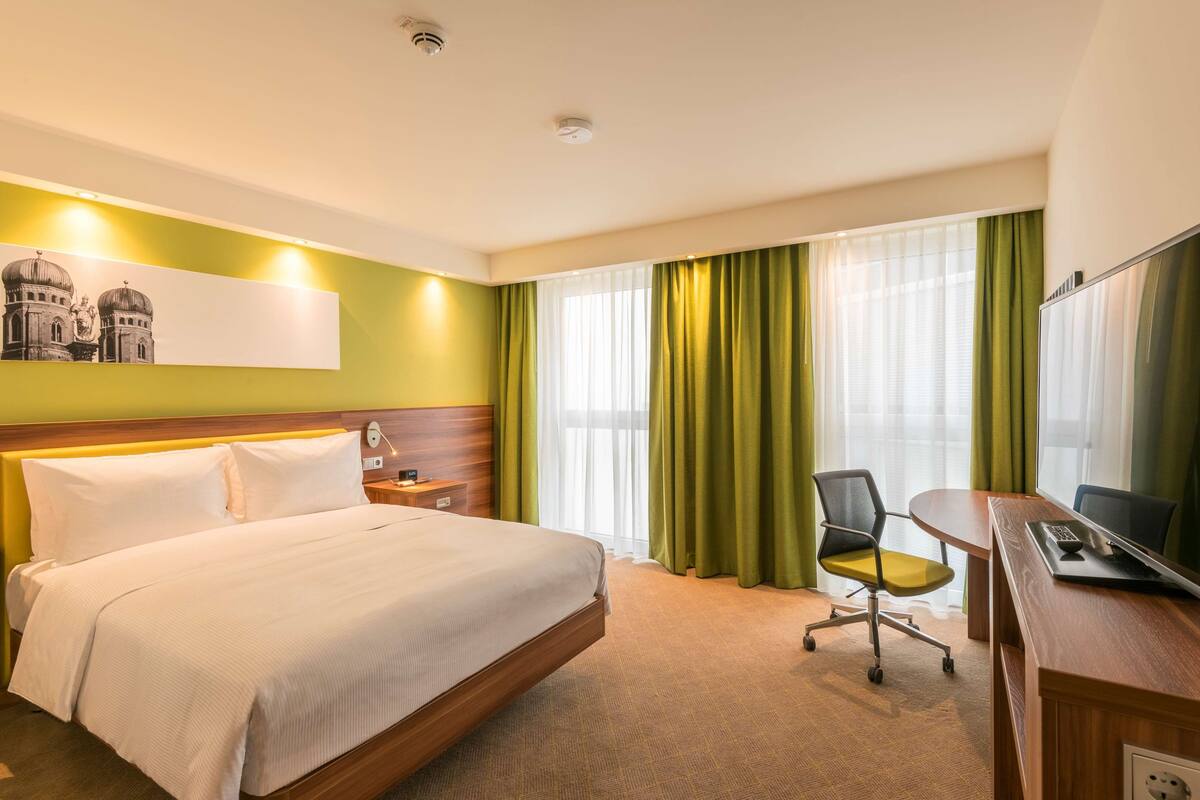 Une chambre d’hôtel avec un lit, un bureau avec un ordinateur, une chaise, une télévision et une fenêtre avec des rideaux.