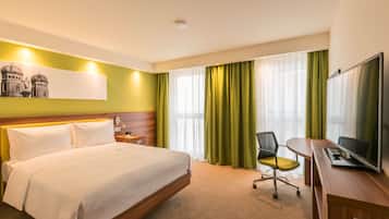 Habitación de hotel con cama, escritorio con computadora, silla, televisor y ventana con cortinas.