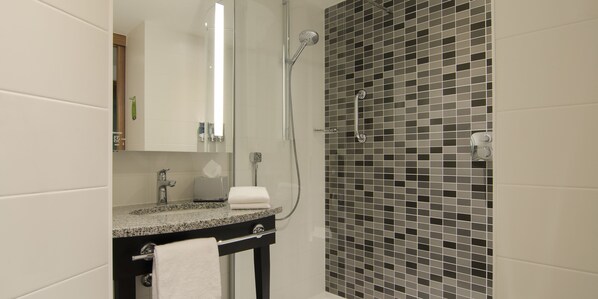 A bathroom with a granite countertop, a mirror, a towel, a shower with a glass door, and a tiled wall. - Hampton by Hilton Munich City West (Munich)