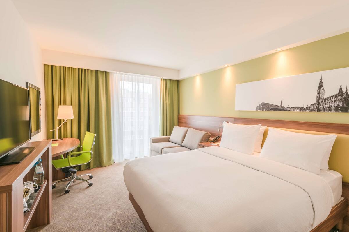 Une chambre d’hôtel avec un lit, un bureau et une chaise, une lampe et une grande fenêtre avec des rideaux.