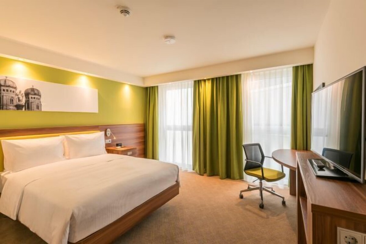 Ein Hotelzimmer mit Bett, Schreibtisch, Stuhl, Fernseher und einem Fenster mit Vorhängen.