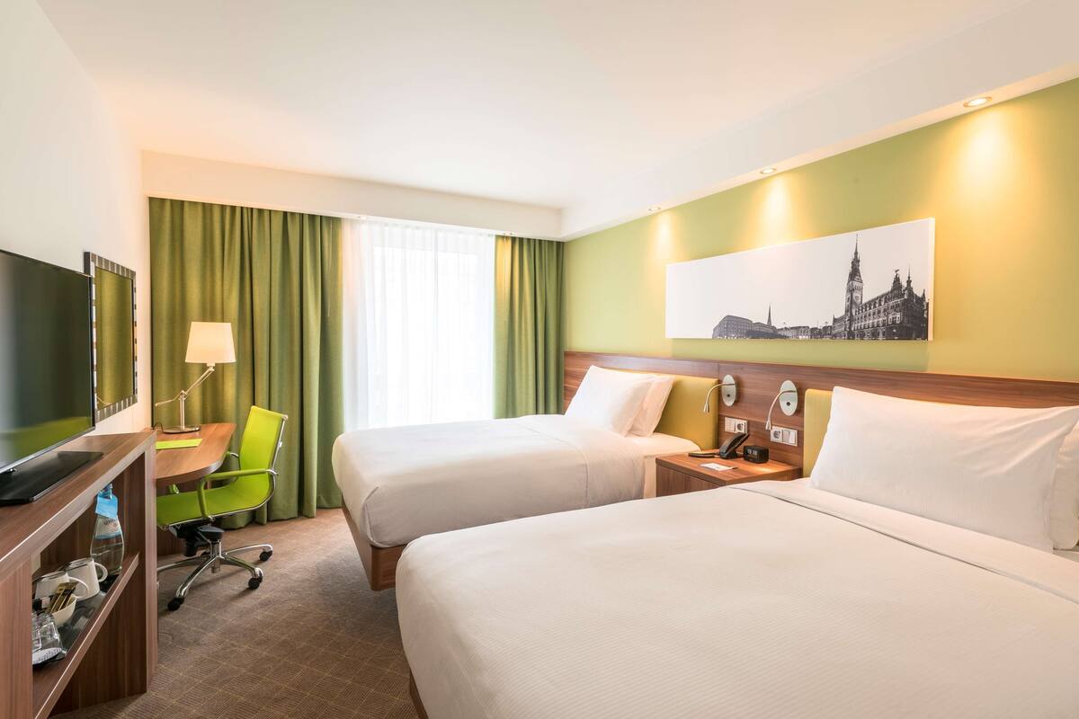 Une chambre d’hôtel avec deux lits, un bureau avec une chaise, une télévision et un tableau au mur.