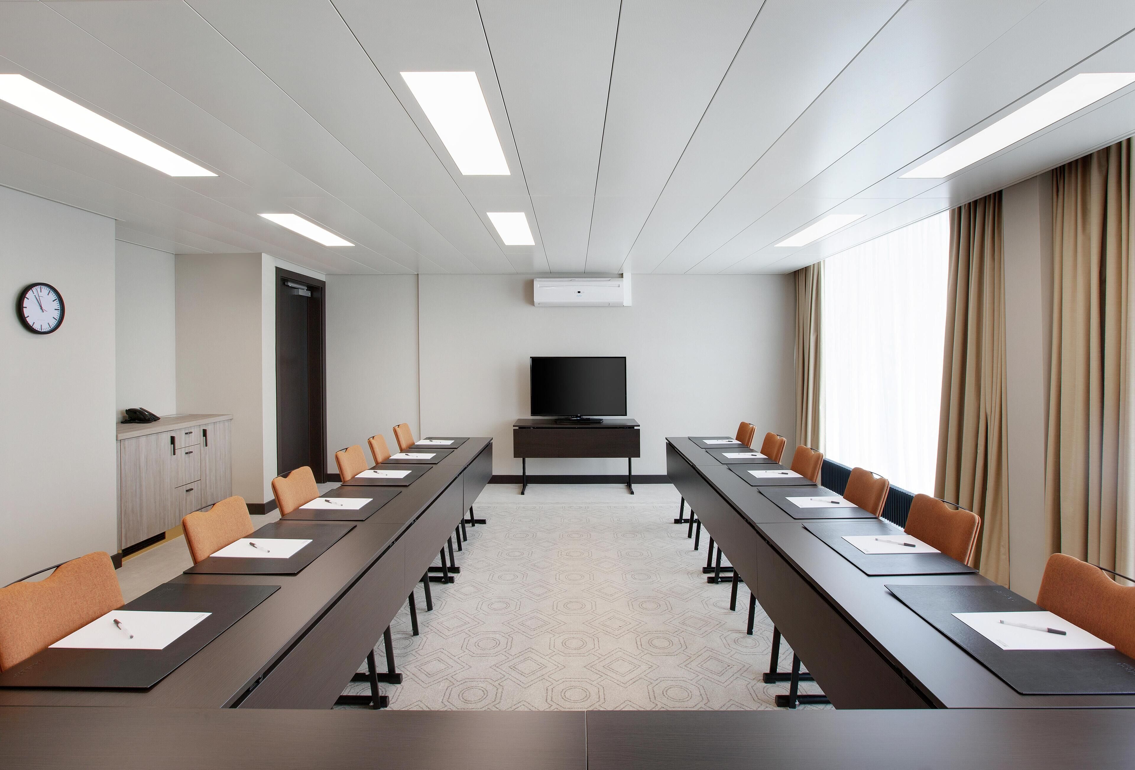 A conference room with a long table, chairs, a clock, a television, and a window with curtains.