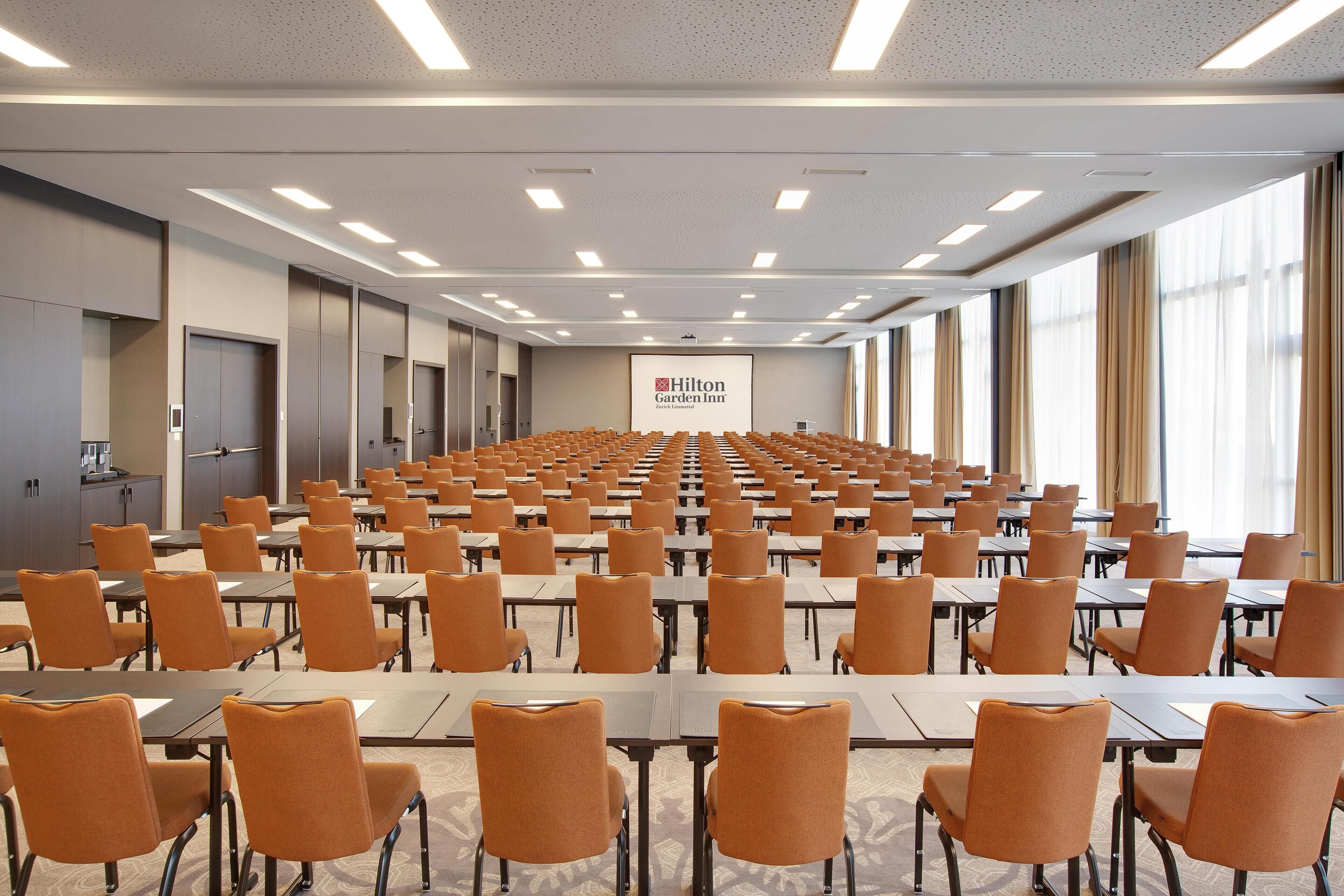 A large conference room with rows of tables and chairs, a podium, and a Hilton Garden Inn sign.