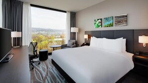 A hotel room with a large bed, a desk with a computer, a chair, a small table, and a view of a cityscape through large windows. - Hilton Garden Inn Zurich Limmattal (Spreitenbach)