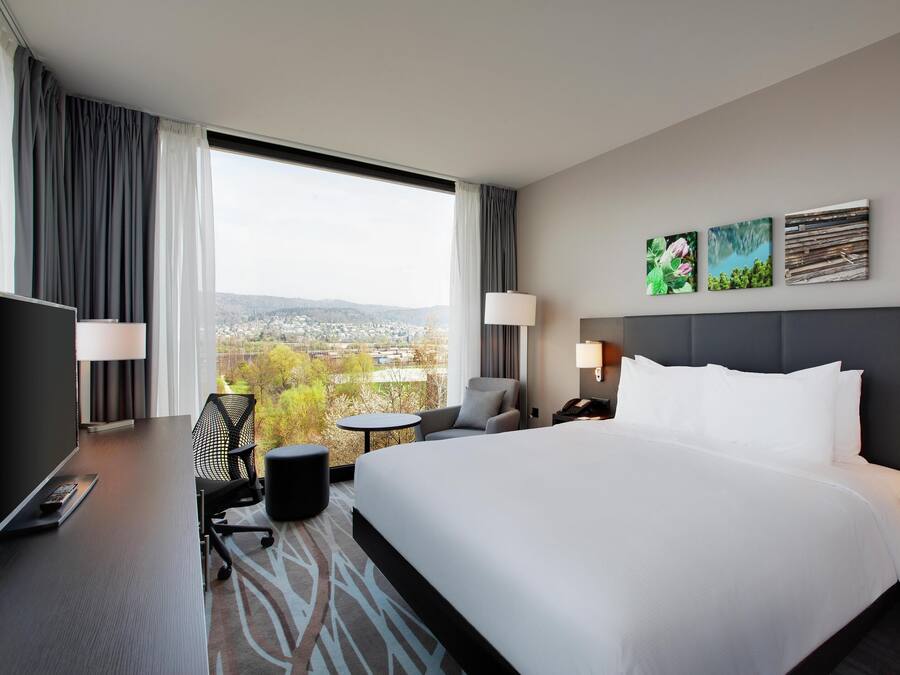 Ein Hotelzimmer mit einem großen Bett, einem Schreibtisch mit Computer, einem Stuhl, einem kleinen Tisch und Blick auf eine Stadtlandschaft durch große Fenster.