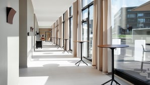 A modern corridor with multiple tables and chairs, large windows, and a view of buildings outside. - Hilton Garden Inn Zurich Limmattal (Spreitenbach)