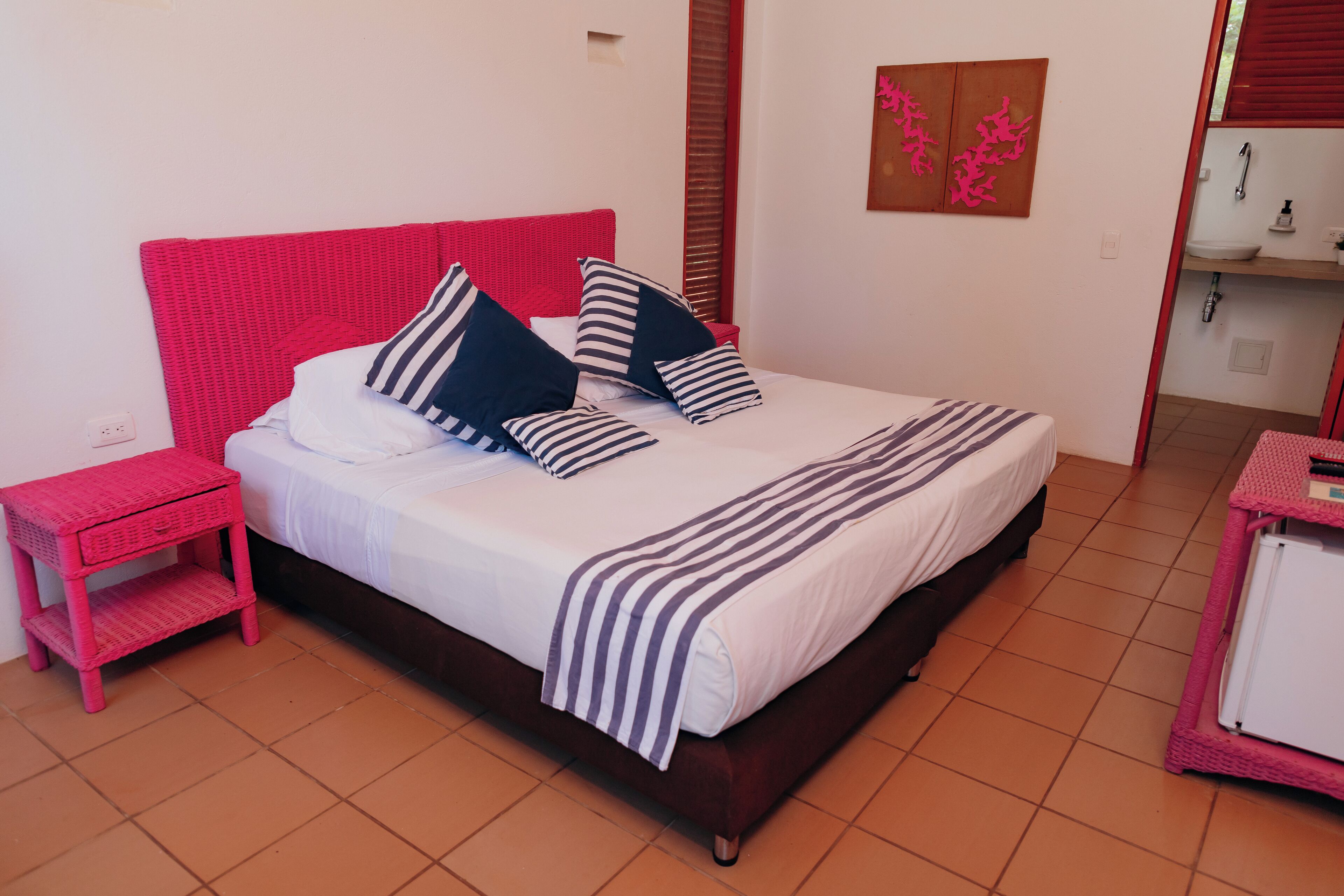 Een net opgemaakt bed met gestreept beddengoed, een rood hoofdeinde en een roze nachtkastje met lade.