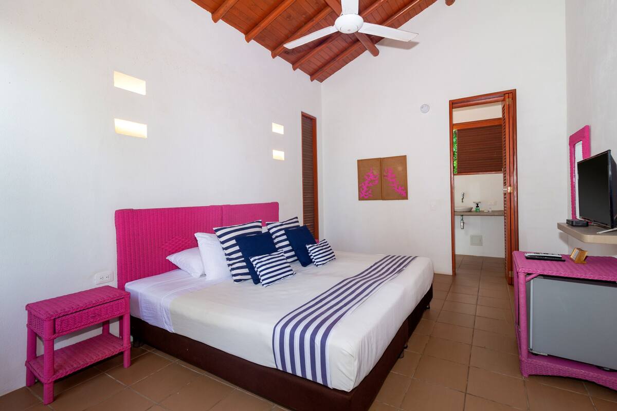 A bedroom with a bed, a pink nightstand, a TV on a pink stand, and a bathroom visible through an open door.