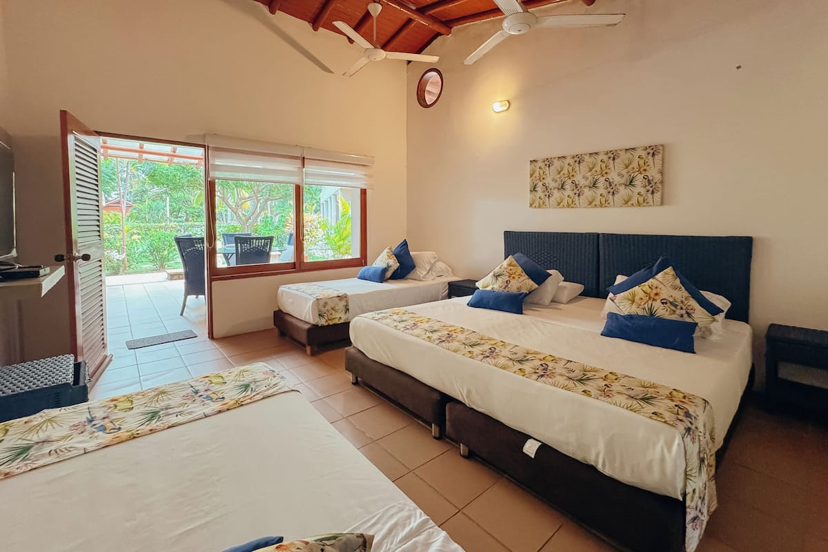 A hotel room with two beds, a ceiling fan, a window with a view of greenery, and a wall-mounted light.
