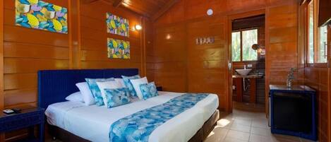 A wooden cabin bedroom with a bed, nightstands, a ceiling fan, artwork, a bathroom with a sink and mirror, and a small table.