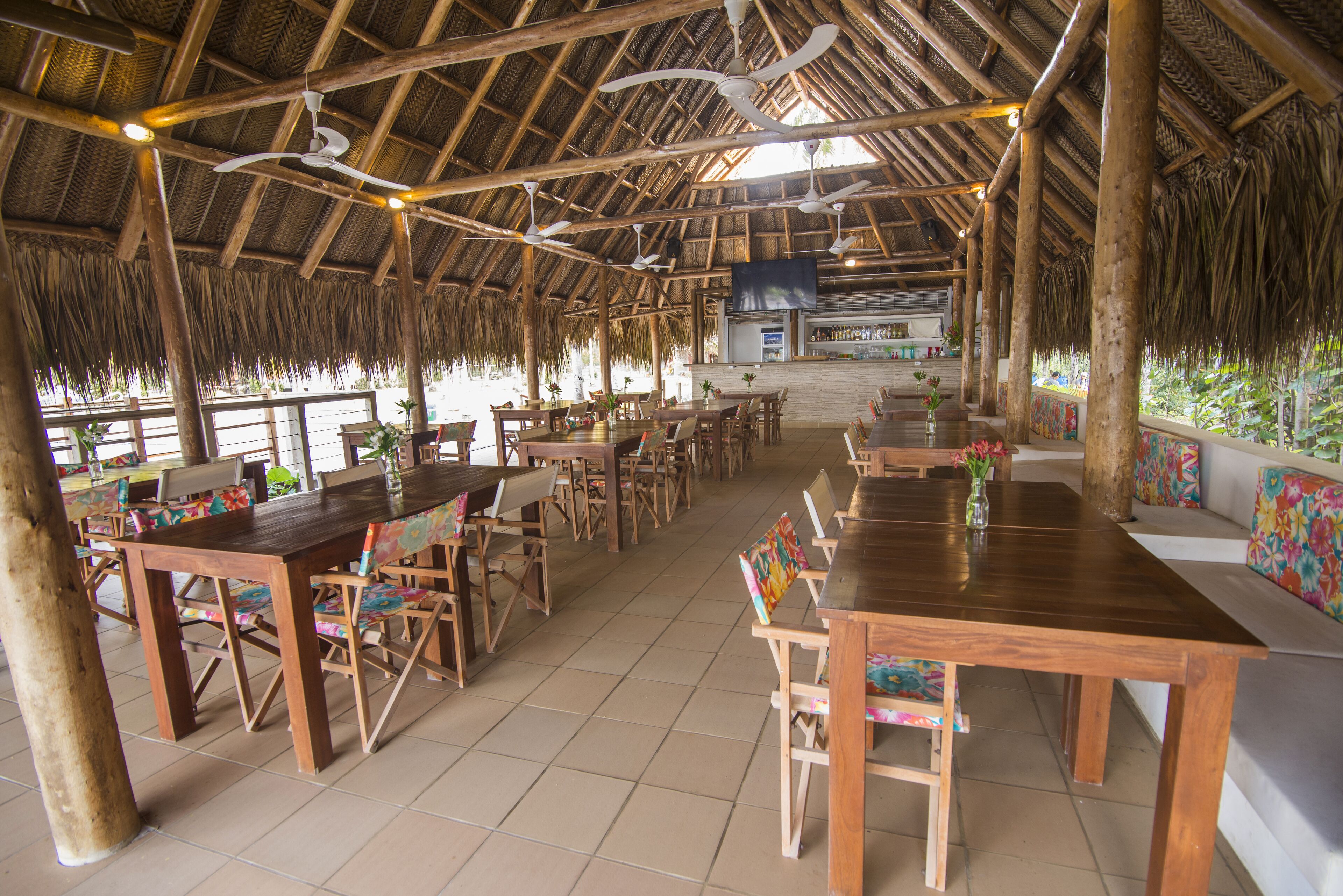 A spacious outdoor dining area with wooden tables and chairs, a thatched roof, and a bar in the background.