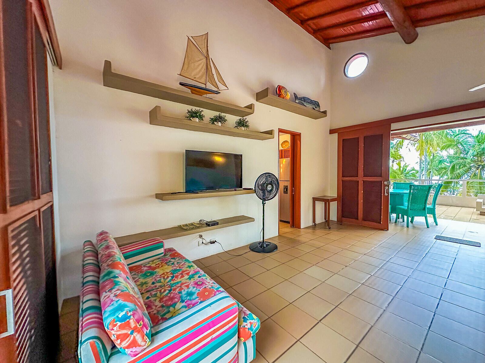 A living room with a sofa, TV, and fan, a balcony with chairs, and a sailboat decoration.