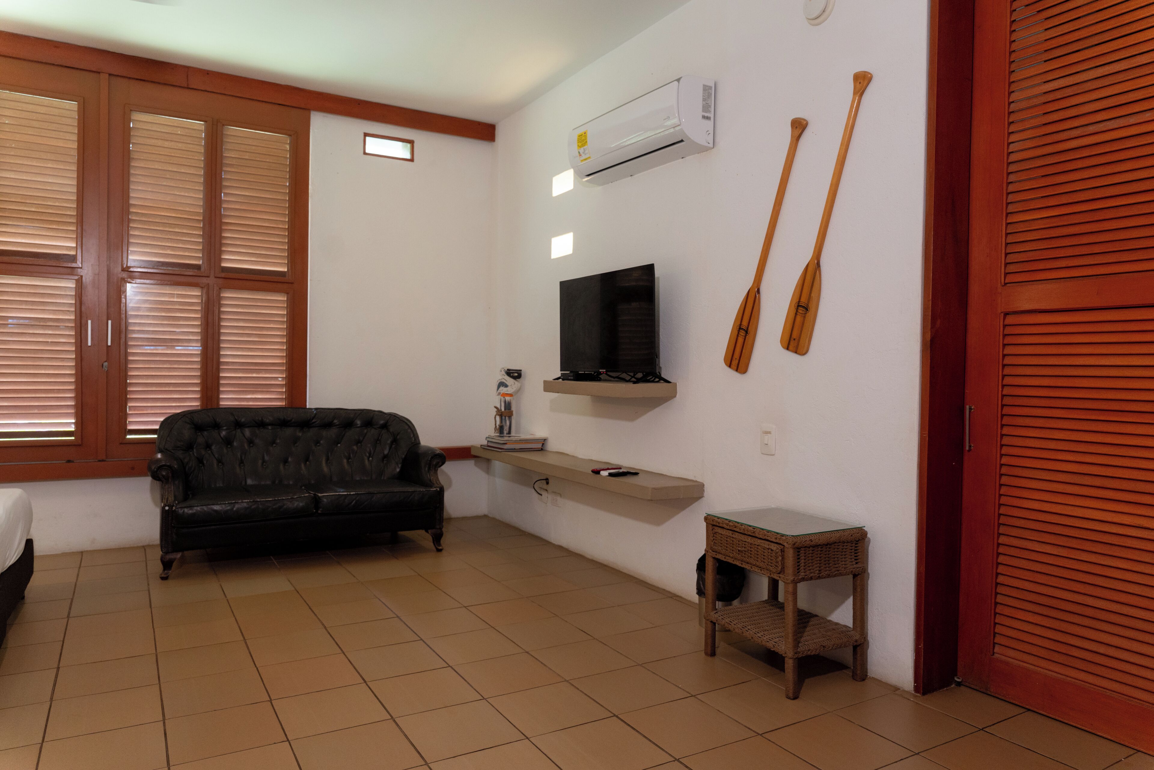 Une pièce avec un canapé noir, un téléviseur fixé au mur, des pagaies en bois, une petite table et une fenêtre avec des stores.