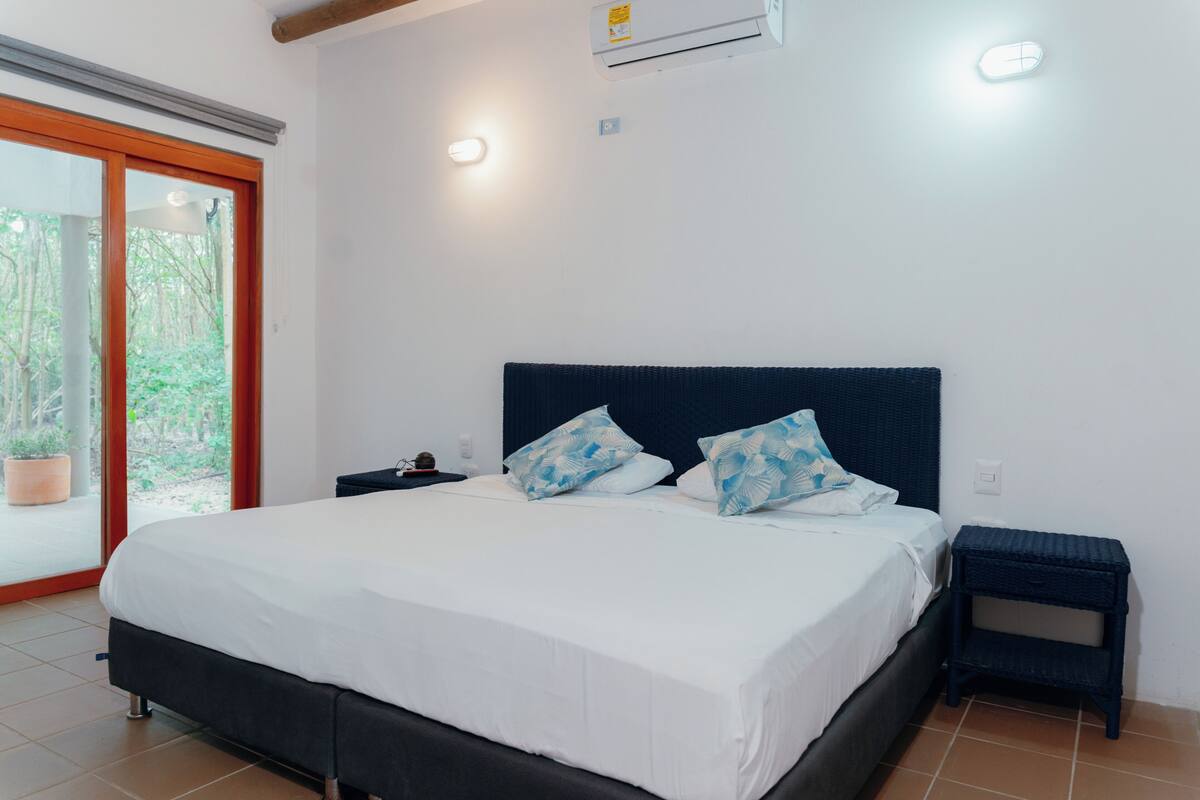 A bedroom with a bed, a nightstand, a window with a view of greenery, and an air conditioning unit.