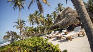 Un resort frente a la playa con cabañas de techo de paja, sillas de descanso y palmeras.