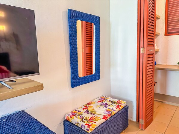 Ein Zimmer mit Fernseher auf einem Schrank, einer Bank mit Blumenkissen, einem blauen gewebten Spiegel und einer Holztür.