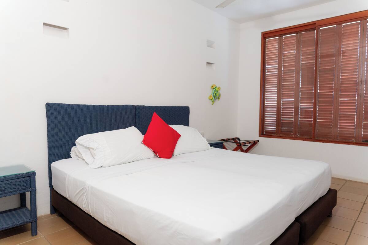 A bed with white bedding and a red pillow, a small side table, and a window with wooden blinds.