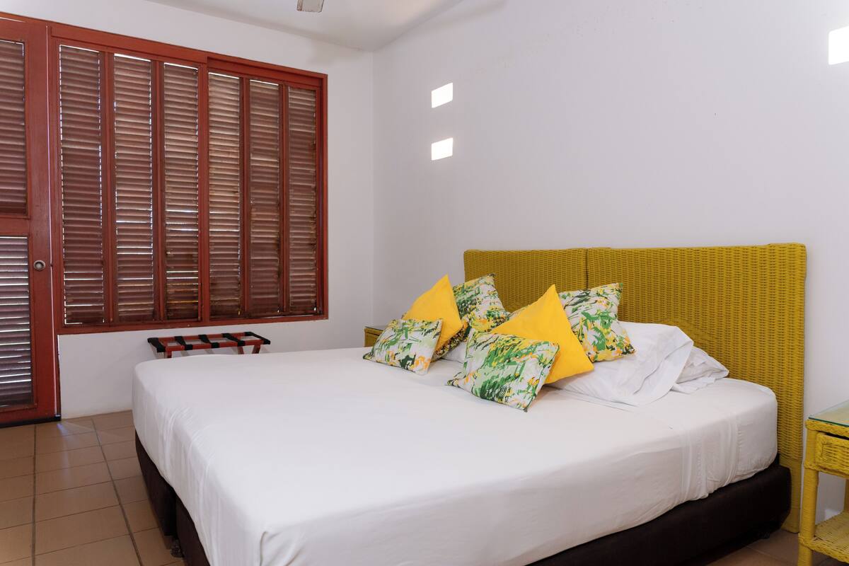 A bedroom with a bed, pillows, a nightstand, a window with blinds, and a door.
