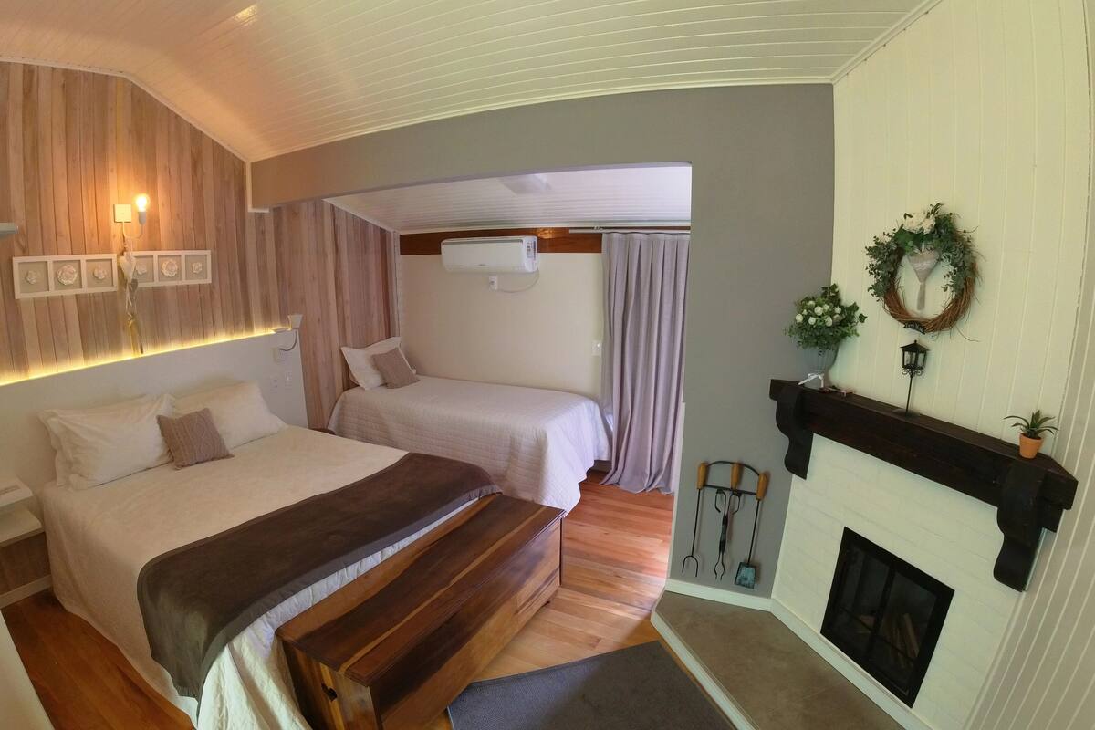 A cozy bedroom with a large bed, a smaller bed, a bench, a fireplace, and a wall-mounted air conditioner.