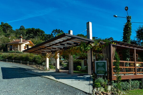 A building with a covered entrance, a sign indicating a reception area, and a view of a house in the background. - Serra Bela Eco Pousada (Urubici)